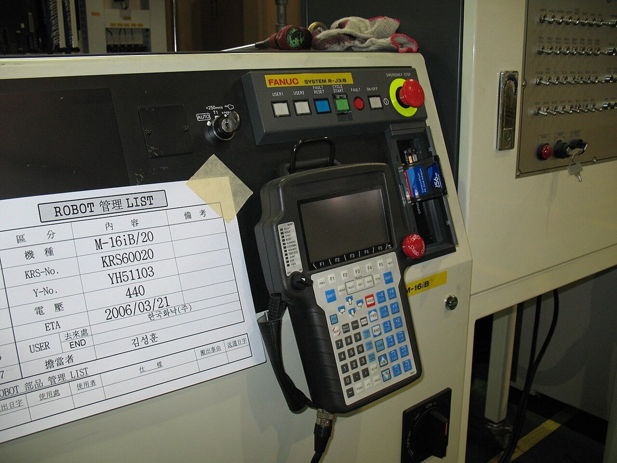 FANUC 제어 캐비닛 앞에 화면과 여러 버튼이 있는 휴대형 조작기가 걸려 있고 위쪽에 빨간 비상 정지 버튼이 보인다.